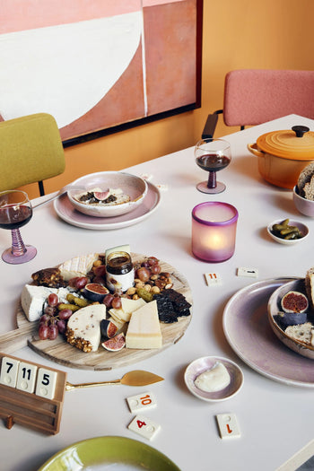 A cozy table for two is set with HKLIVING Chef Ceramics: Deep Plate M, assorted cheeses, grapes, figs, bread, pickles, red wine glasses, a lit candle, a board game, and colorful decor in a warm room.
