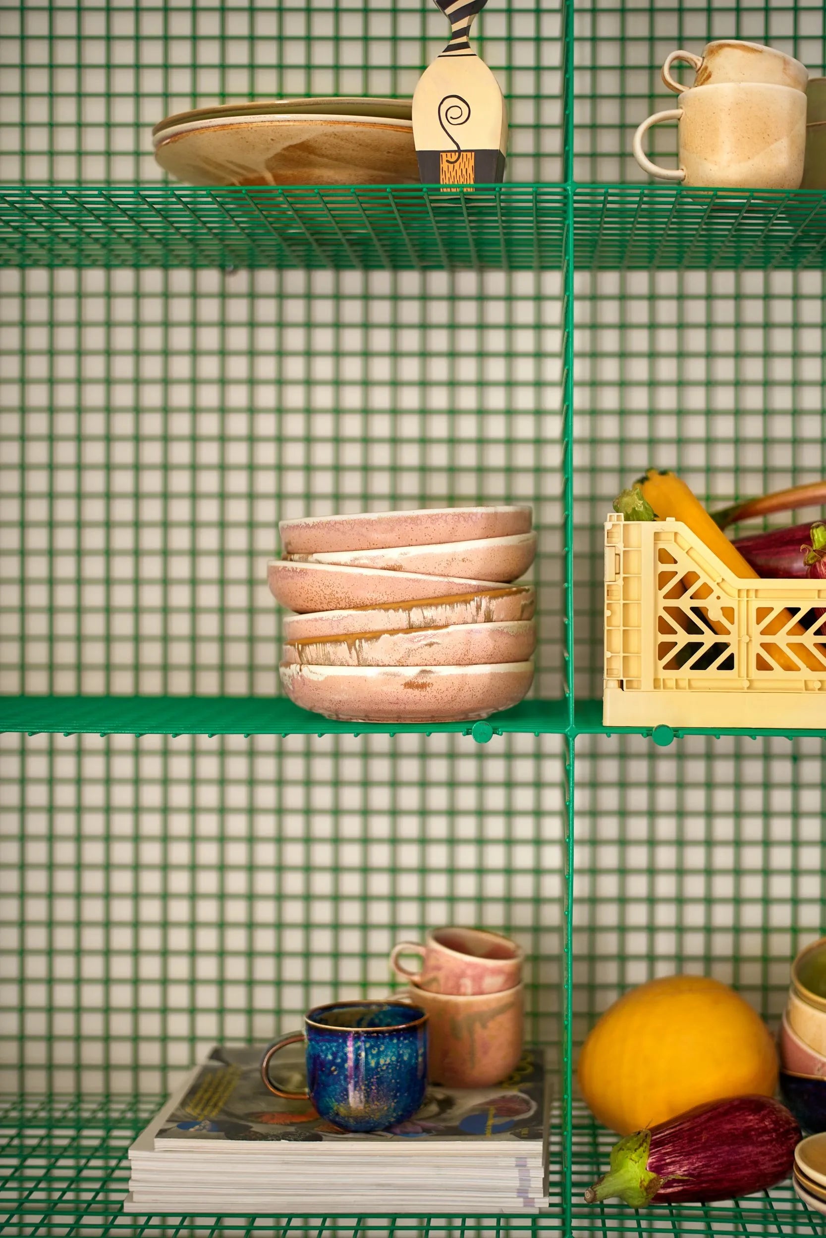 Shelves display hand-glazed HKLIVING Chef Ceramics, including the Deep Plate M, bowls, and cups, alongside a crate of vegetables, a yellow melon, a decorative plate, and magazines—all set against a green grid-patterned backdrop.