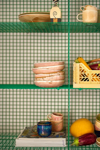 Shelves display hand-glazed HKLIVING Chef Ceramics, including the Deep Plate M, bowls, and cups, alongside a crate of vegetables, a yellow melon, a decorative plate, and magazines—all set against a green grid-patterned backdrop.