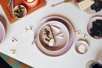 An overhead shot of a table featuring HKLIVING Chef Ceramics: Deep Plate M, cheese, cake with cream and a strawberry, pickles, game tiles, and the book "Pâtisserie.