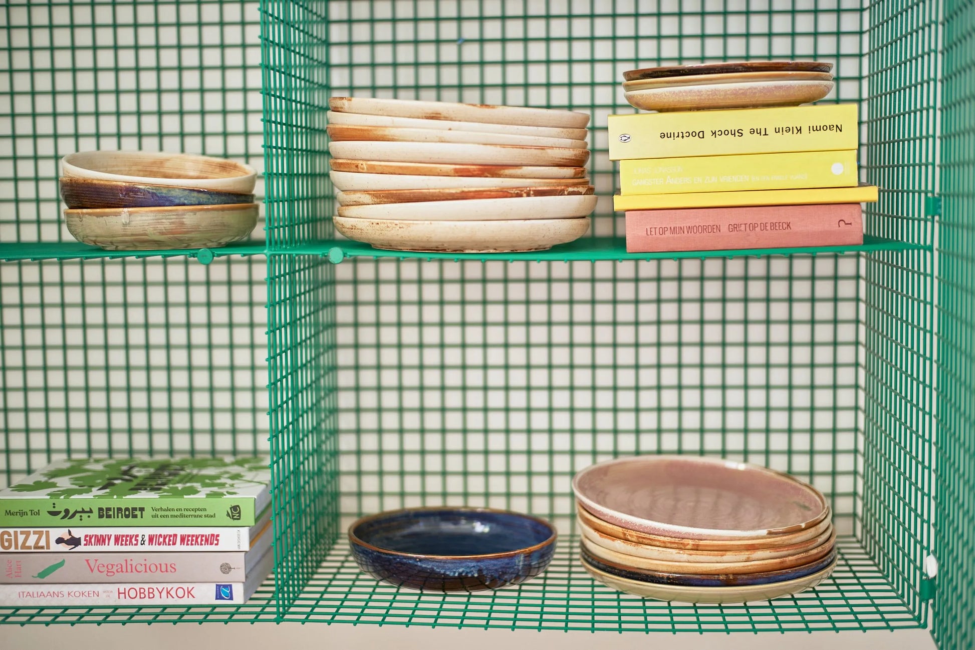 A green wire shelf displays stacks of HKLIVING Chef Ceramics: Dinner Plates, including a rustic pink artisanal-glazed plate by HKLIVING, with bowls on middle and bottom shelves and colorful cookbooks on the bottom left.