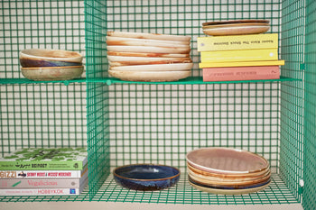 A green wire shelf displays stacks of HKLIVING Chef Ceramics: Dinner Plates, including a rustic pink artisanal-glazed plate by HKLIVING, with bowls on middle and bottom shelves and colorful cookbooks on the bottom left.