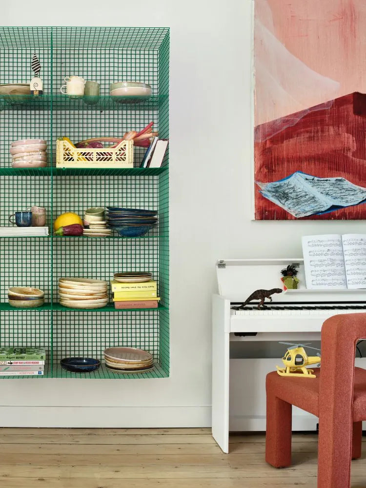 A green wire wall shelf displays stacked ceramics, including the HKLIVING Chef Ceramics: Dinner Plate in pink, bowls, mugs, and books. Nearby are a white piano with sheet music and toys, a red chair, and an abstract painting.