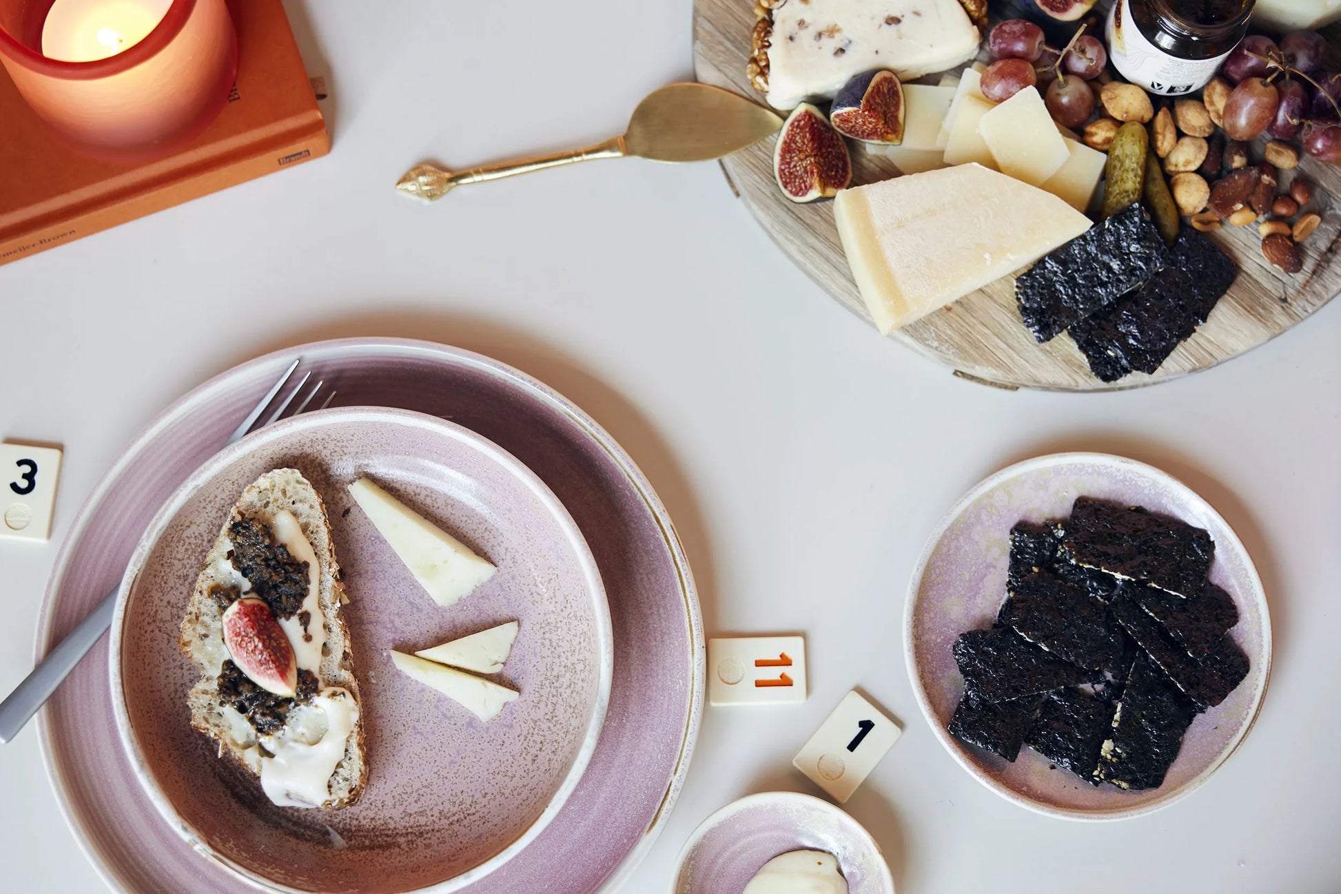 A table set with the HKLIVING Chef Ceramics: Dinner Plate, cheese, crackers topped with figs, seaweed, nuts, a cheese knife, a candle, and number tiles on a white surface.