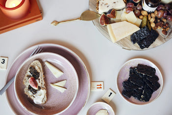 A table set with the HKLIVING Chef Ceramics: Dinner Plate, cheese, crackers topped with figs, seaweed, nuts, a cheese knife, a candle, and number tiles on a white surface.