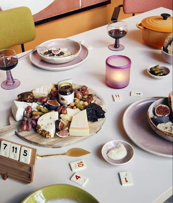 A meal table with cheese, grapes, figs, bread on a wooden platter, red wine glasses, a lit candle, a yellow pot, Rummikub tiles scattered around, and an HKLIVING Chef Ceramics: Dinner Plate by HKLIVING with artisanal glaze.