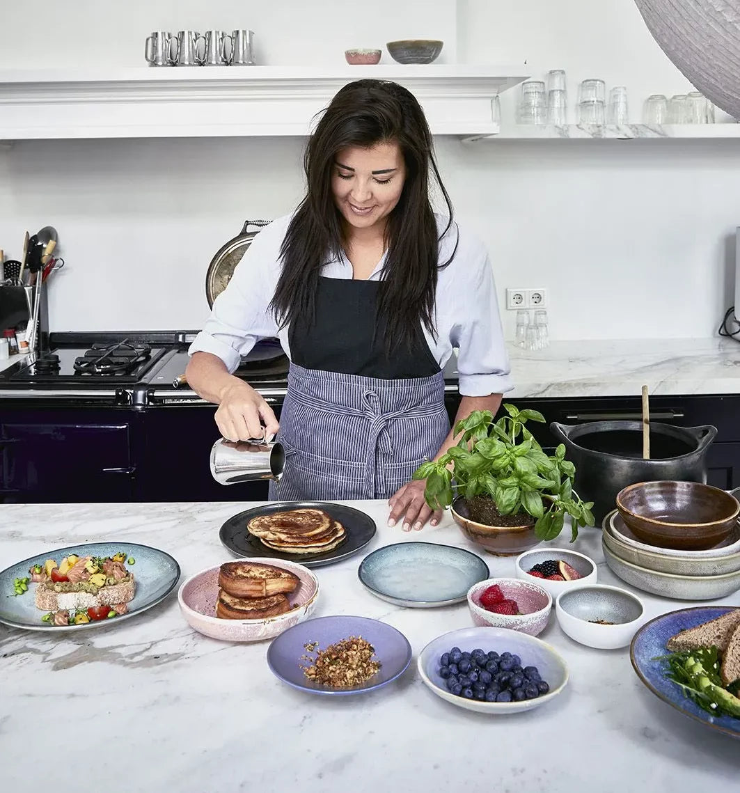 A woman in an apron pours syrup on pancakes in a bright kitchen, with plates of fresh fruit, granola, and herbs on a marble countertop. Berries are served in the HKLIVING Chef Ceramics: Flat Bowl by HKLIVING.