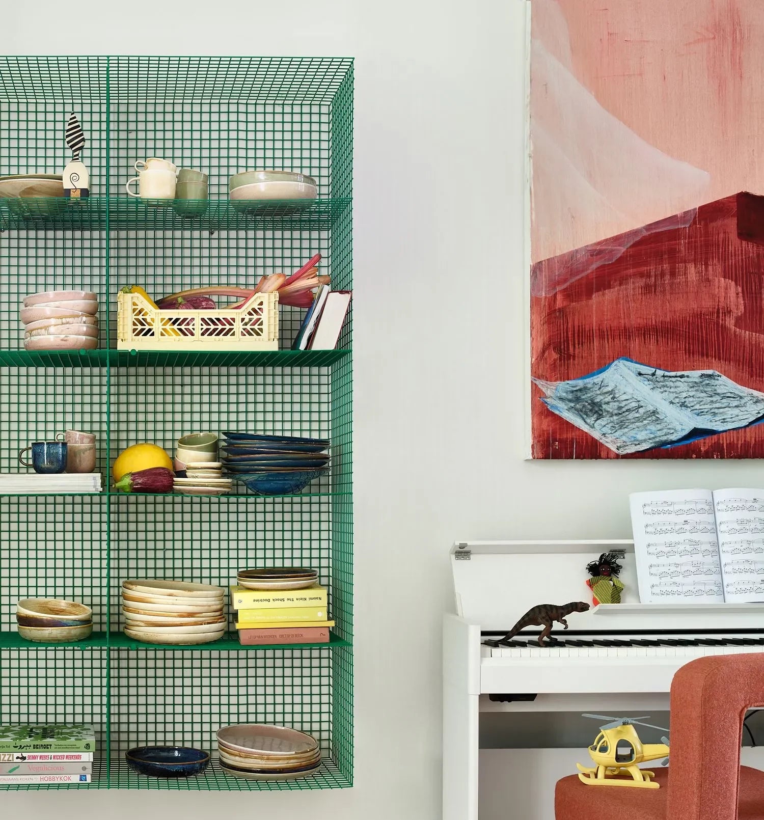 A green wire wall shelf displays HKLIVING Chef Ceramics Mugs, a yellow basket, and fruit. Next to it is a piano with sheet music, a toy dinosaur, and an orange chair. Above hangs an abstract red and blue painting.