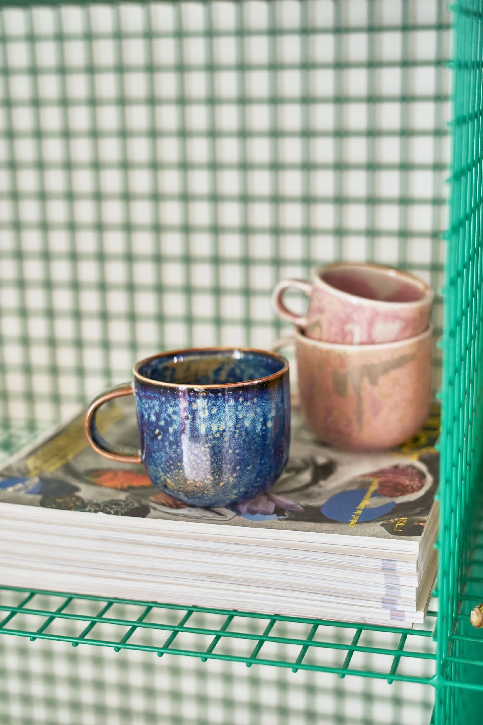 Two HKLIVING Chef Ceramics mugs—a blue speckled mug and two stacked pink mugs—rest atop magazines on a green wire shelf with a green and white checkered background.