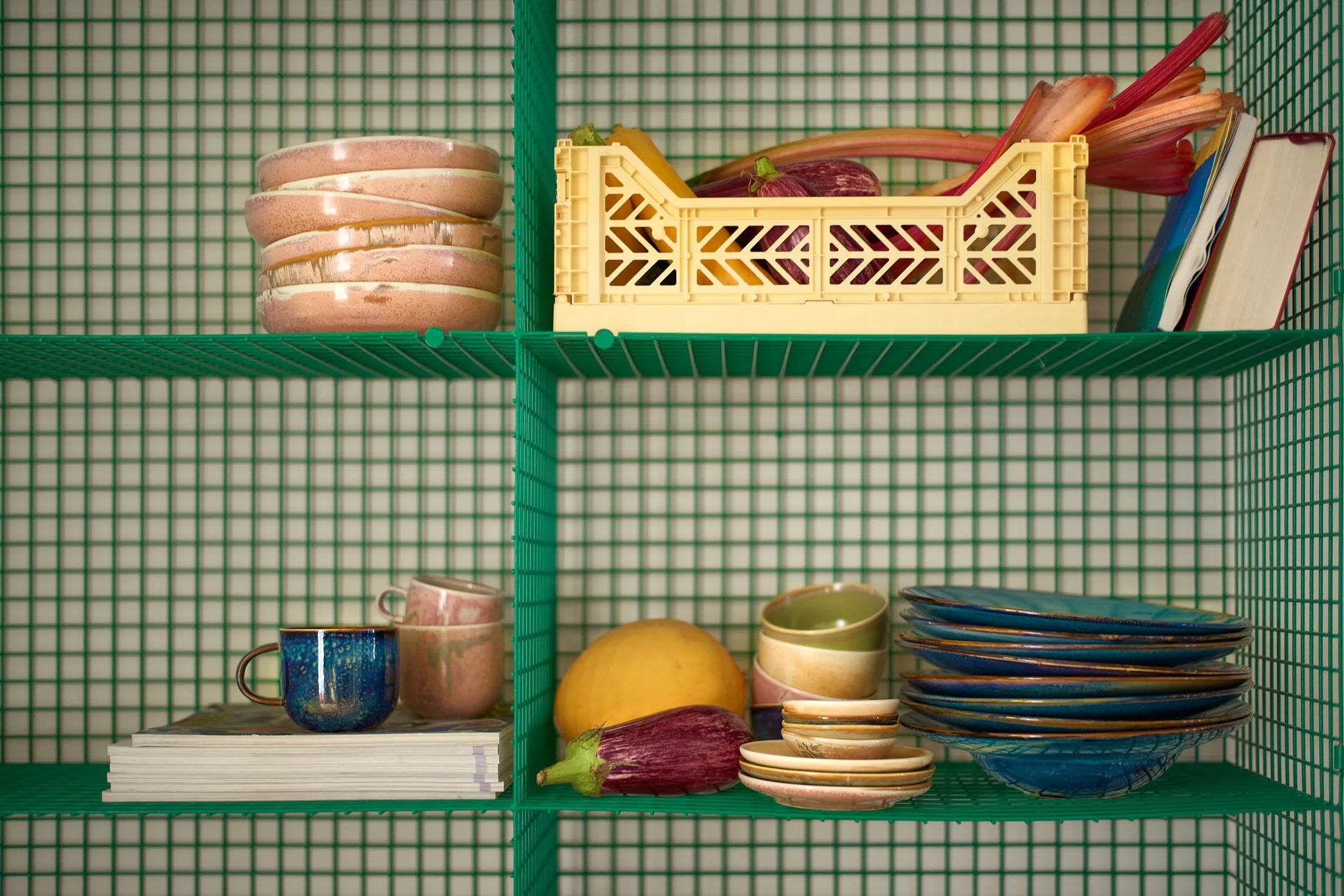 Green wire shelving holds stacked dishes, cups, the HKLIVING Chef Ceramics: Mug by HKLIVING, a yellow crate with vegetables, a book, yellow fruit, and two eggplants—all set against a grid-patterned background.