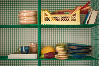 A green wire shelf holds stacked bowls, plates, cups, and the HKLIVING Chef Ceramics: Mug by HKLIVING, plus a yellow crate with vegetables, a book, a mango, an eggplant—all set against a checkered background.