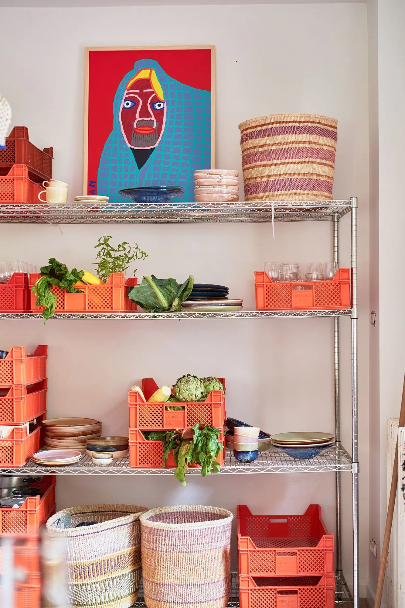 Metal shelves with orange crates display HKLIVING Chef Ceramics: Pasta Plate and fresh vegetables. Woven baskets are on the top and bottom shelves, and a colorful portrait with a blue background hangs above.