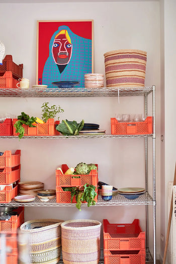 Metal shelves with orange crates display HKLIVING Chef Ceramics: Pasta Plate and fresh vegetables. Woven baskets are on the top and bottom shelves, and a colorful portrait with a blue background hangs above.