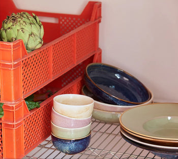 A red plastic crate holds an artichoke and leafy greens. Next to it, HKLIVING Chef Ceramics: Pasta Plate and handcrafted ceramic bowls in assorted colors are stacked on a metal wire shelf.
