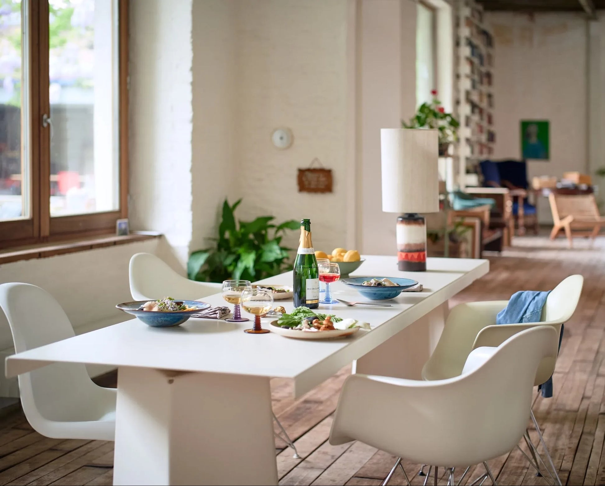 A spacious, bright room with wooden floors and large windows features a modern dining table set for a meal with HKLIVING Chef Ceramics: Pasta Plates, white chairs, and lush green plants.