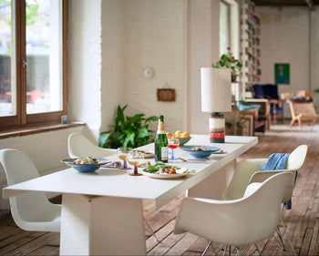 A spacious, bright room with wooden floors and large windows features a modern dining table set for a meal with HKLIVING Chef Ceramics: Pasta Plates, white chairs, and lush green plants.