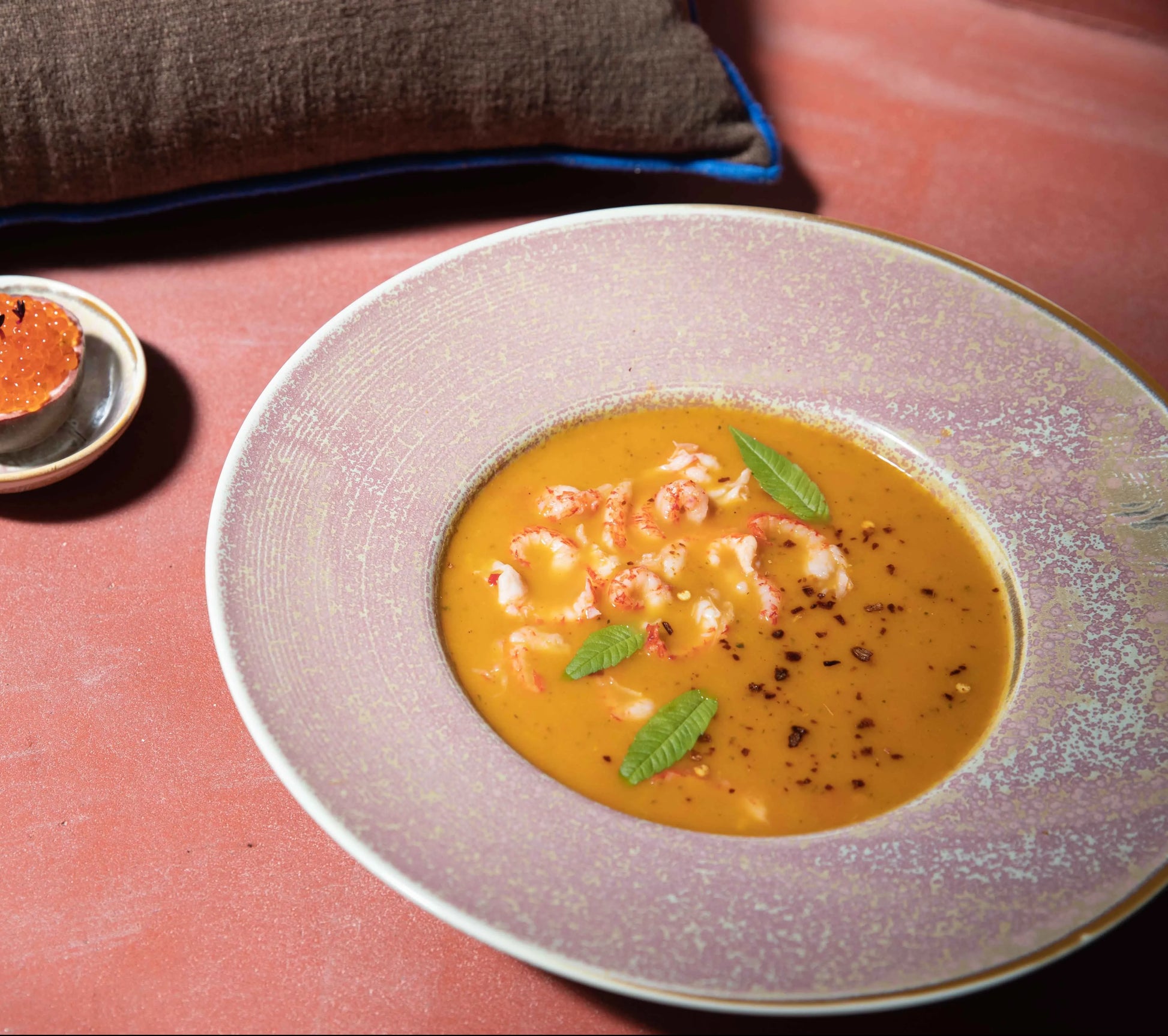 A creamy orange soup with herbs and seafood is served in an HKLIVING Chef Ceramics: Pasta Plate by HKLIVING, placed on a pink surface with a brown napkin and a small round appetizer on the side.