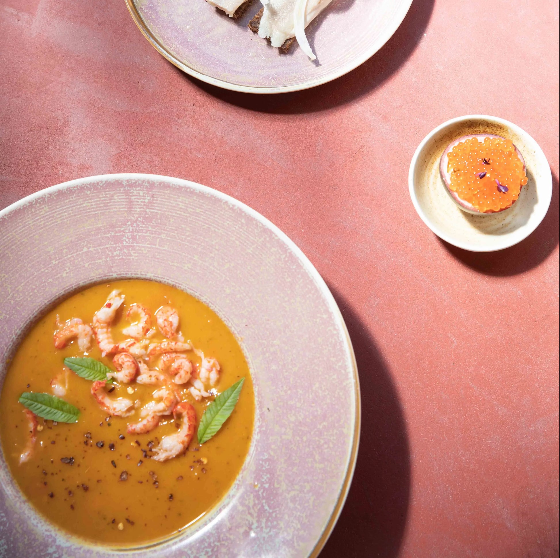 A creamy orange soup garnished with herbs and shrimp is served in an HKLIVING Chef Ceramics Pasta Plate, alongside bread, fish, and a dish of orange caviar, all arranged on a pinkish-red tabletop.