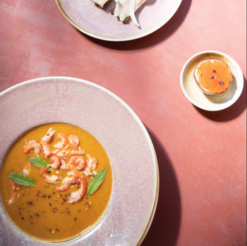 A creamy orange soup garnished with herbs and shrimp is served in an HKLIVING Chef Ceramics Pasta Plate, alongside bread, fish, and a dish of orange caviar, all arranged on a pinkish-red tabletop.