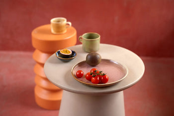 A HKLIVING Chef Ceramics: Side Plate with cherry tomatoes and a dark fruit rests on a round beige table, beside cups and a saucer with half a passion fruit—each piece crafted by HKLIVING from hand-glazed chef ceramics.