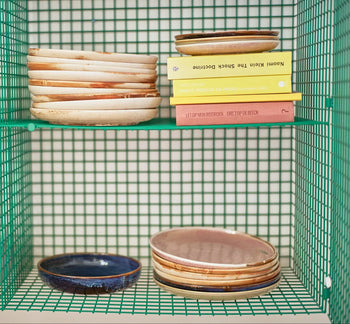A green wire shelf displays stacks of HKLIVING Chef Ceramics: Side Plates in earthy tones, with three yellow and green books neatly arranged on the top shelf beside the plates.