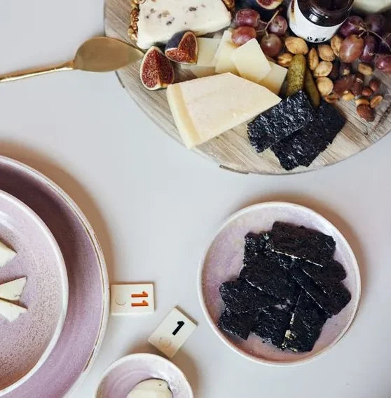A HKLIVING Chef Ceramics: Side Plate in pink holds sliced cheese and seaweed, beside a round wooden board with cheese, figs, grapes, nuts, pickles, a gold knife, and “1” numbered markers on the table.