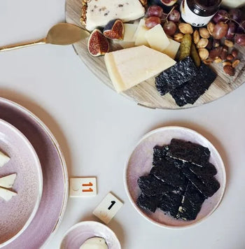 A HKLIVING Chef Ceramics: Side Plate in pink holds sliced cheese and seaweed, beside a round wooden board with cheese, figs, grapes, nuts, pickles, a gold knife, and “1” numbered markers on the table.