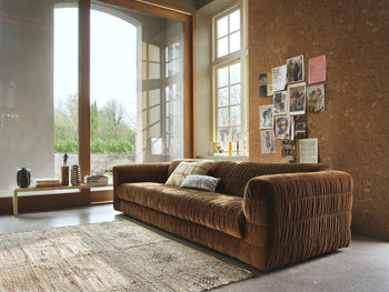 A cozy living room with the HKLIVING Club Couch, featuring pleated upholstery, a textured rug, and a cork wall adorned with photos and postcards. Sunlight pours through tall windows onto books and decor on a low wooden bench.