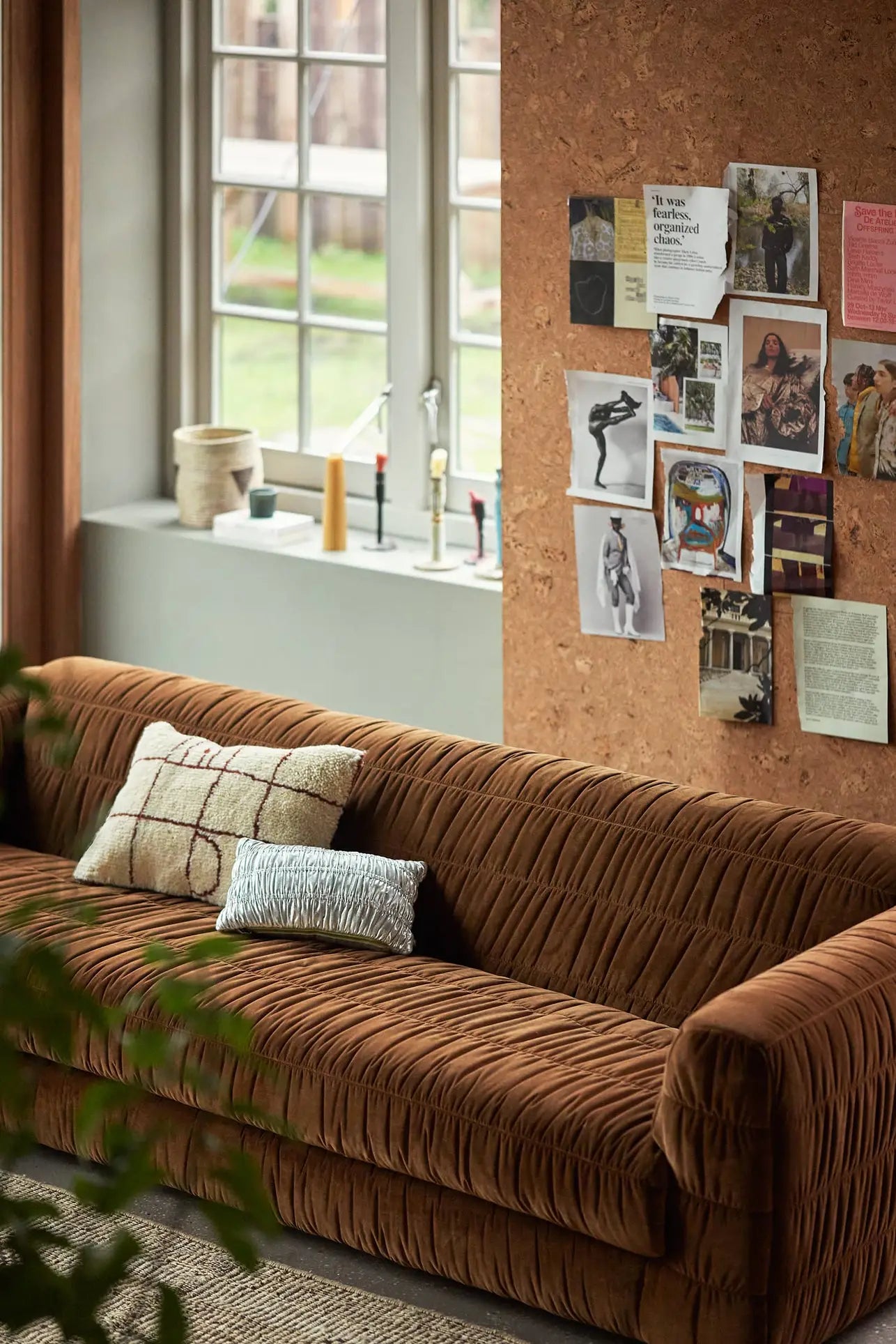 A cozy living room with the HKLIVING Club Couch in brown velvet and pleated upholstery, two pillows, a large window with candles and vases on the sill, and a cork wall decorated with photos—ideal for modern interiors.
