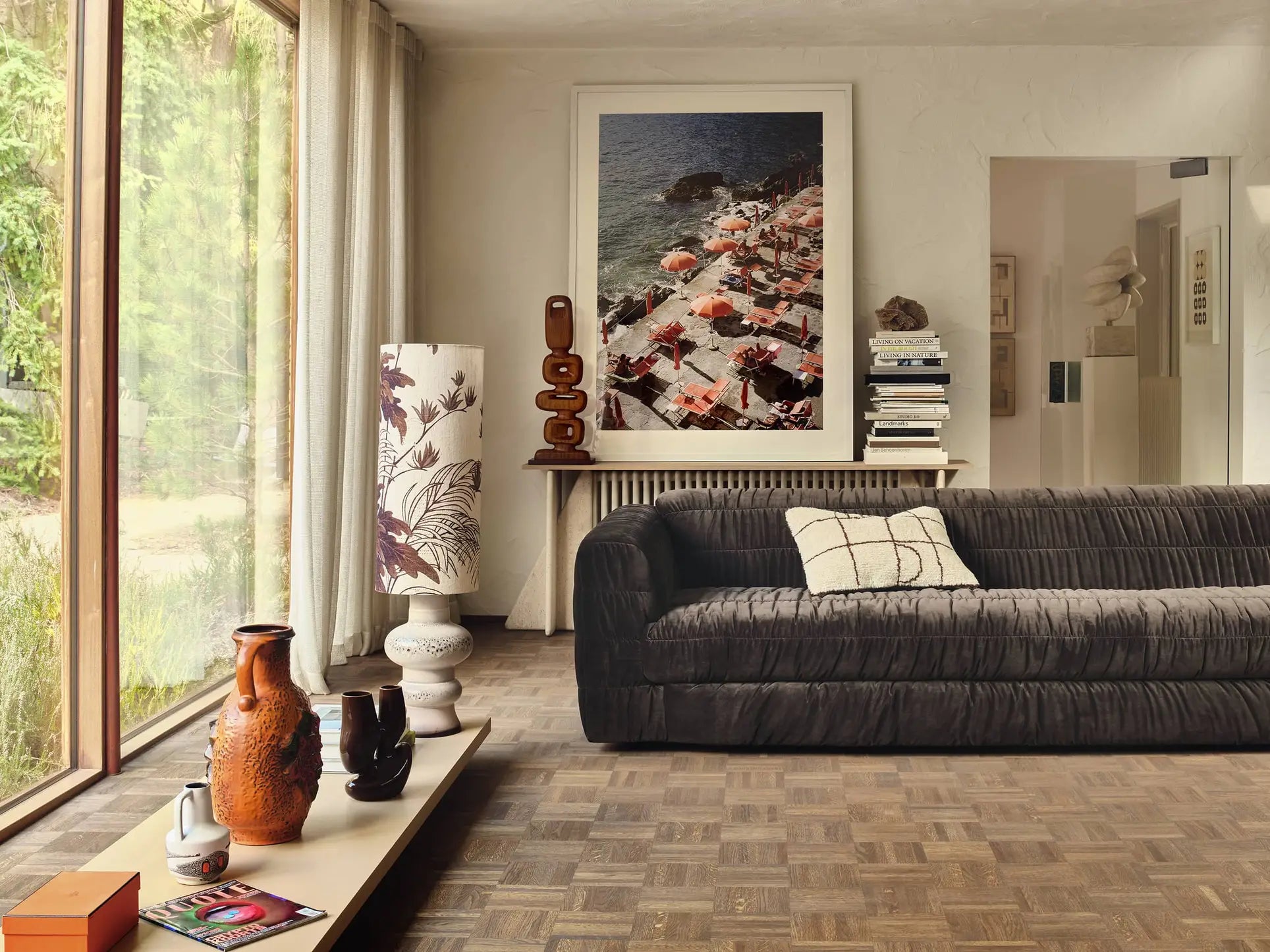 A modern living room features the HKLIVING Club Couch with pleated upholstery, a brown textured sofa, patterned pillows, a tall lamp with floral shade, vases on a low table, stacked books, and a framed aerial beach photo. Sunlight streams in.