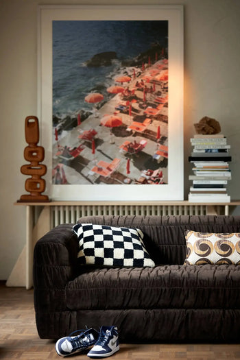 A cozy living room featuring the HKLIVING Club Couch with patterned pillows, sneakers on the floor, a stack of books, a wooden sculpture, and a large framed orange beach photograph on the wall.