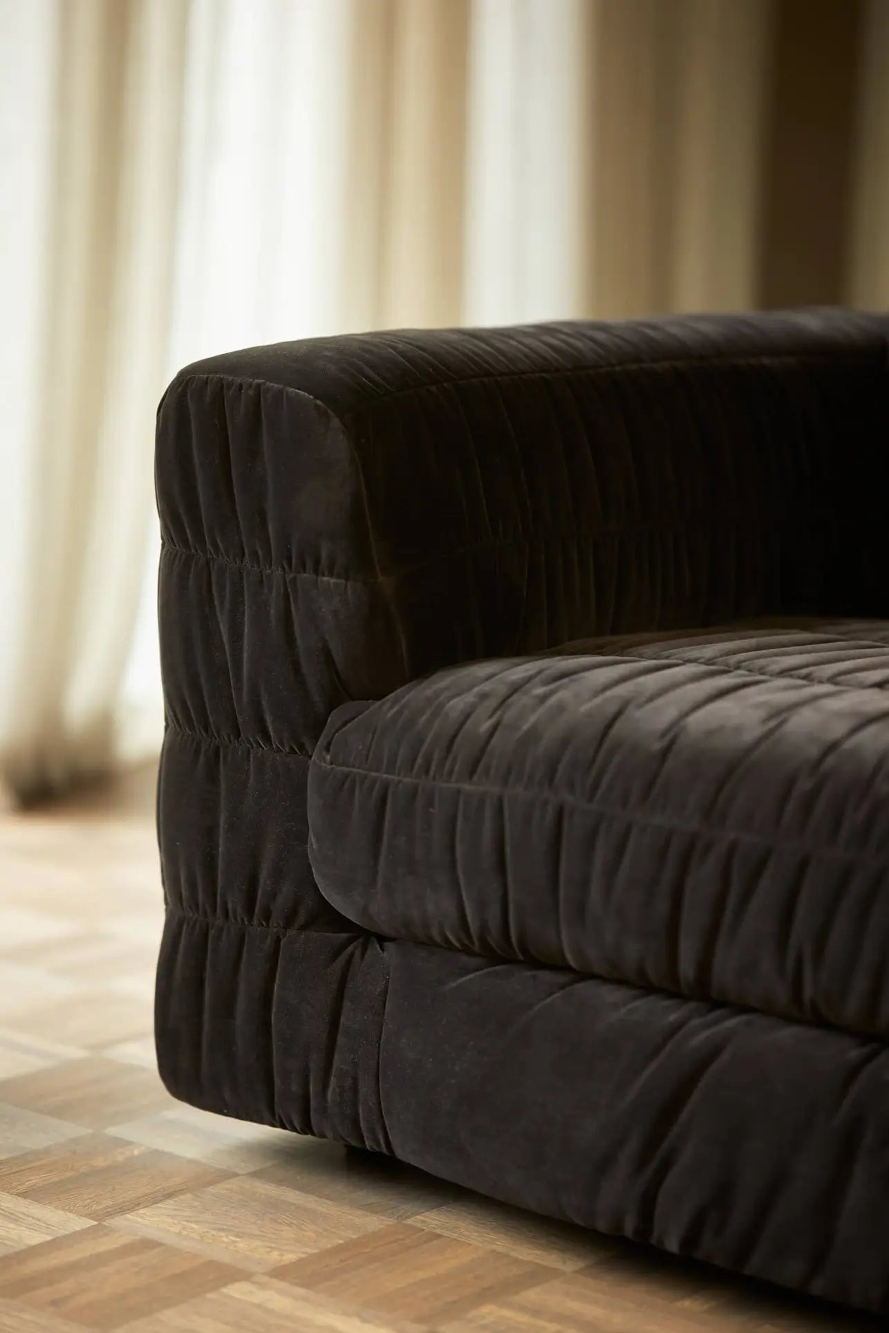 The HKLIVING Club Couch by HKLIVING features dark quilted velvet and wide armrests, set on a wooden parquet floor with softly lit sheer curtains—an elegant addition to modern interiors.