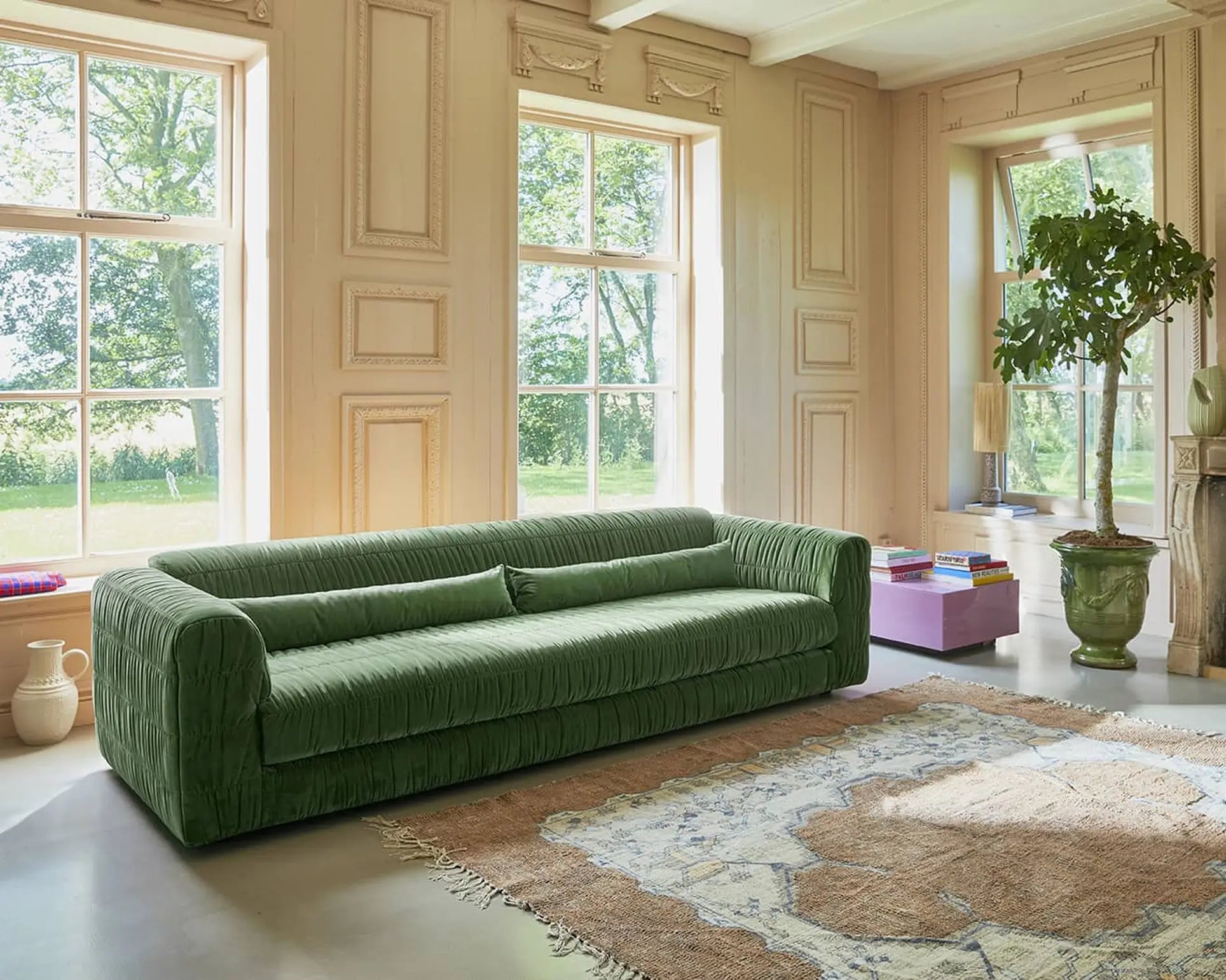 A spacious room with tall windows, beige paneled walls, a green HKLIVING Club Couch, a potted tree, and a pink low table with books sits on a large patterned rug. Sunlight streams in, creating bright modern interiors.