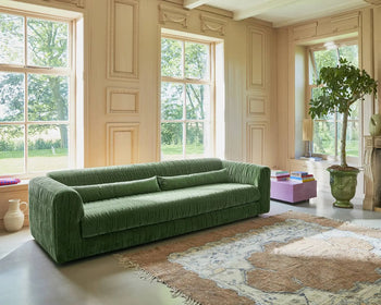 A spacious room with tall windows, beige paneled walls, a green HKLIVING Club Couch, a potted tree, and a pink low table with books sits on a large patterned rug. Sunlight streams in, creating bright modern interiors.