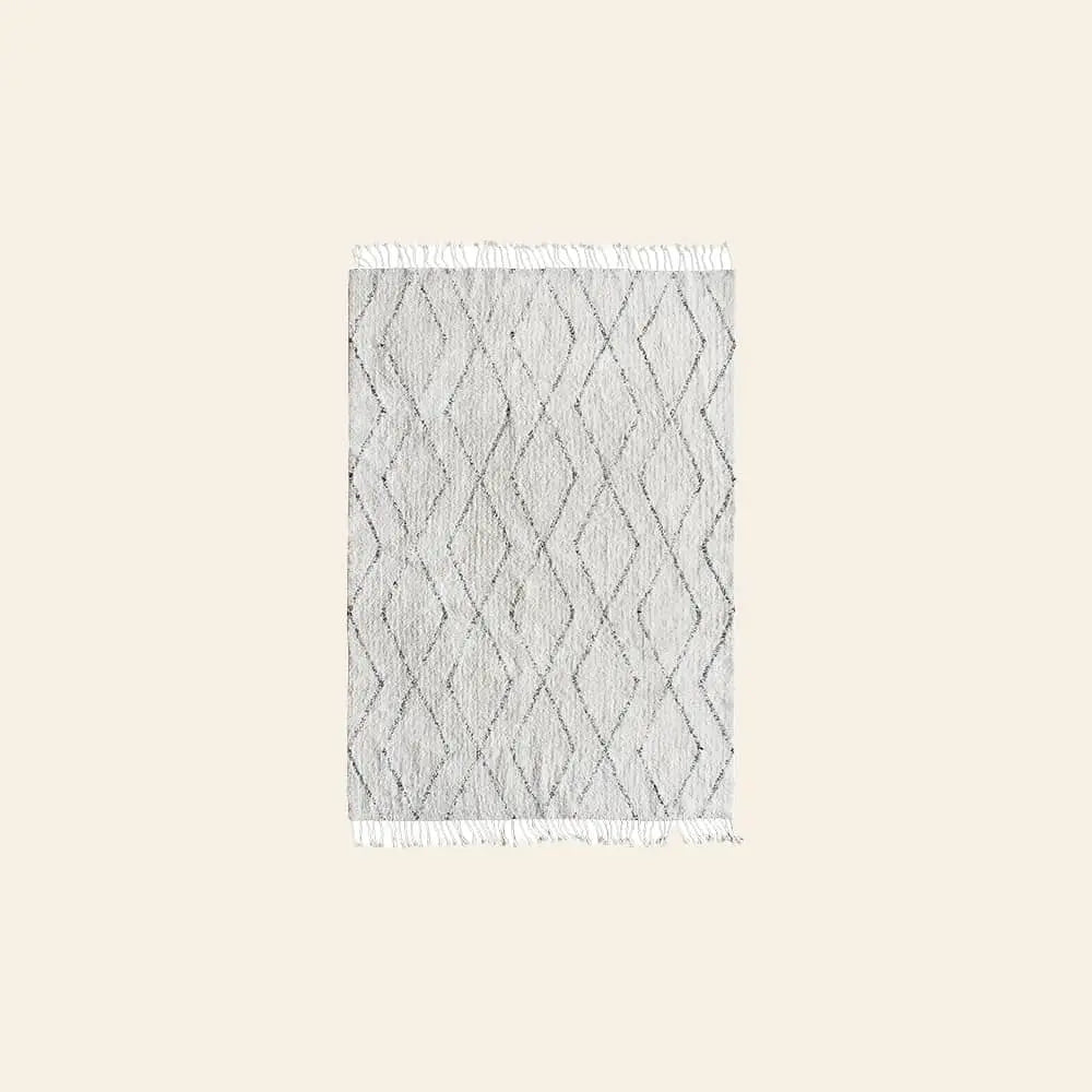 The HKLIVING Cotton Berber Rug (140x200) by HKLIVING is a rectangular, light gray rug with a subtle diamond pattern and white fringe on the short sides, shown against a plain beige background.