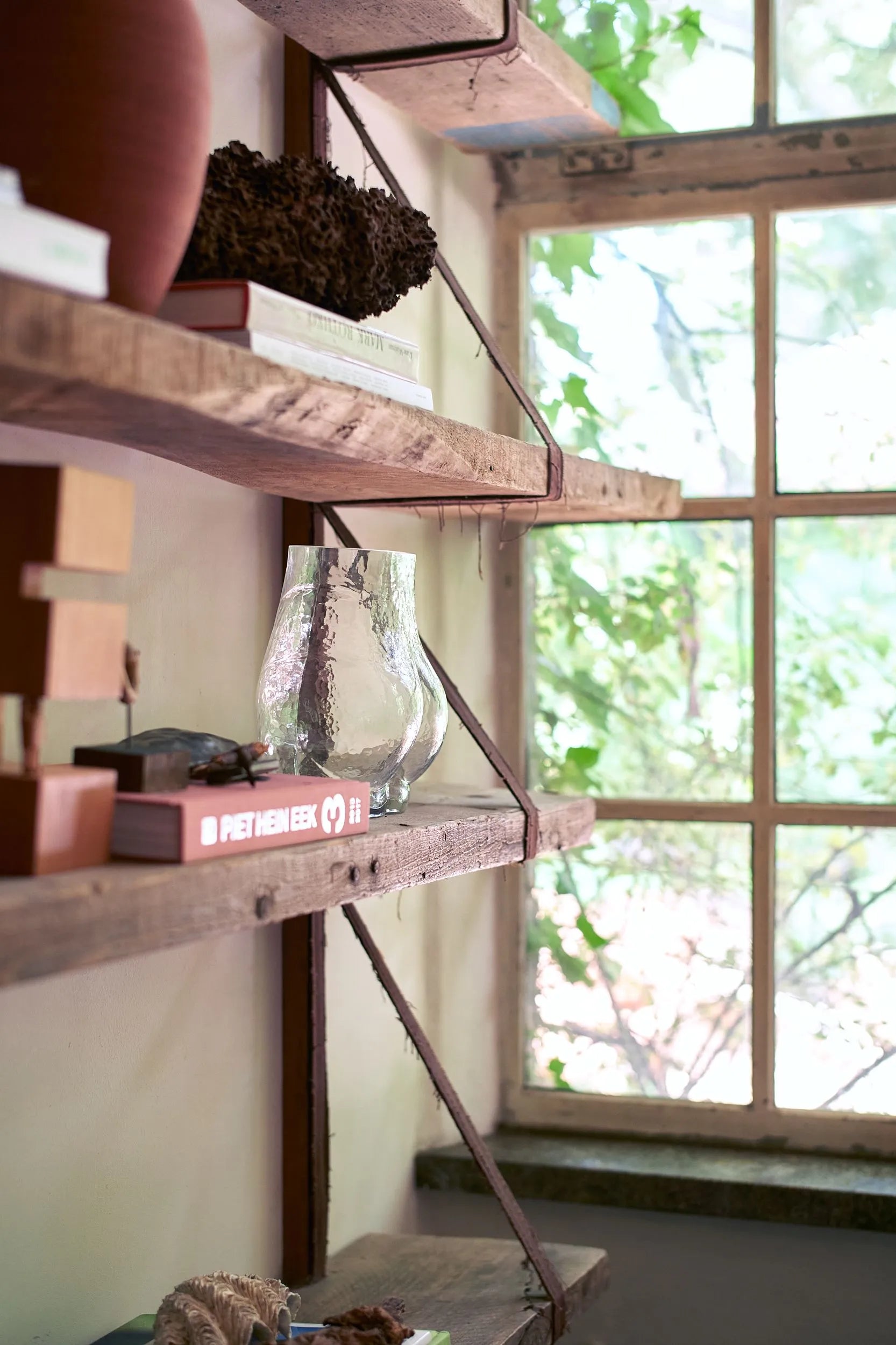 Wooden shelves with books, decor, and the HKLIVING Glass Bum Vase by HKLIVING are mounted on a wall beside a large window. Sunlight pours in, highlighting greenery outside and creating a warm, inviting glow indoors.