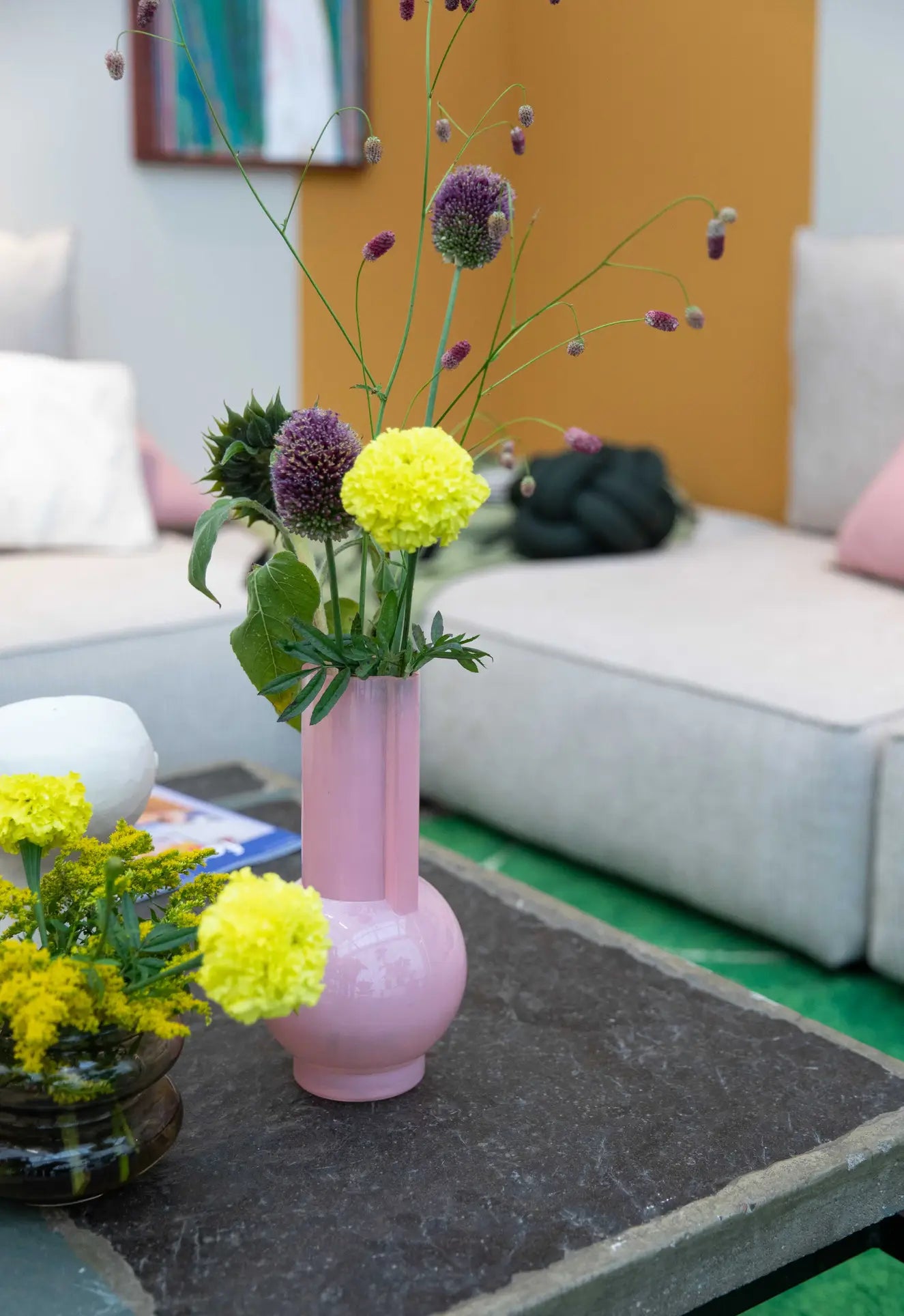 An HKLIVING Glass Vase Flamingo Pink with yellow and purple flowers sits on a dark coffee table beside a smaller yellow flower arrangement, with light sofas and colorful cushions in the background.