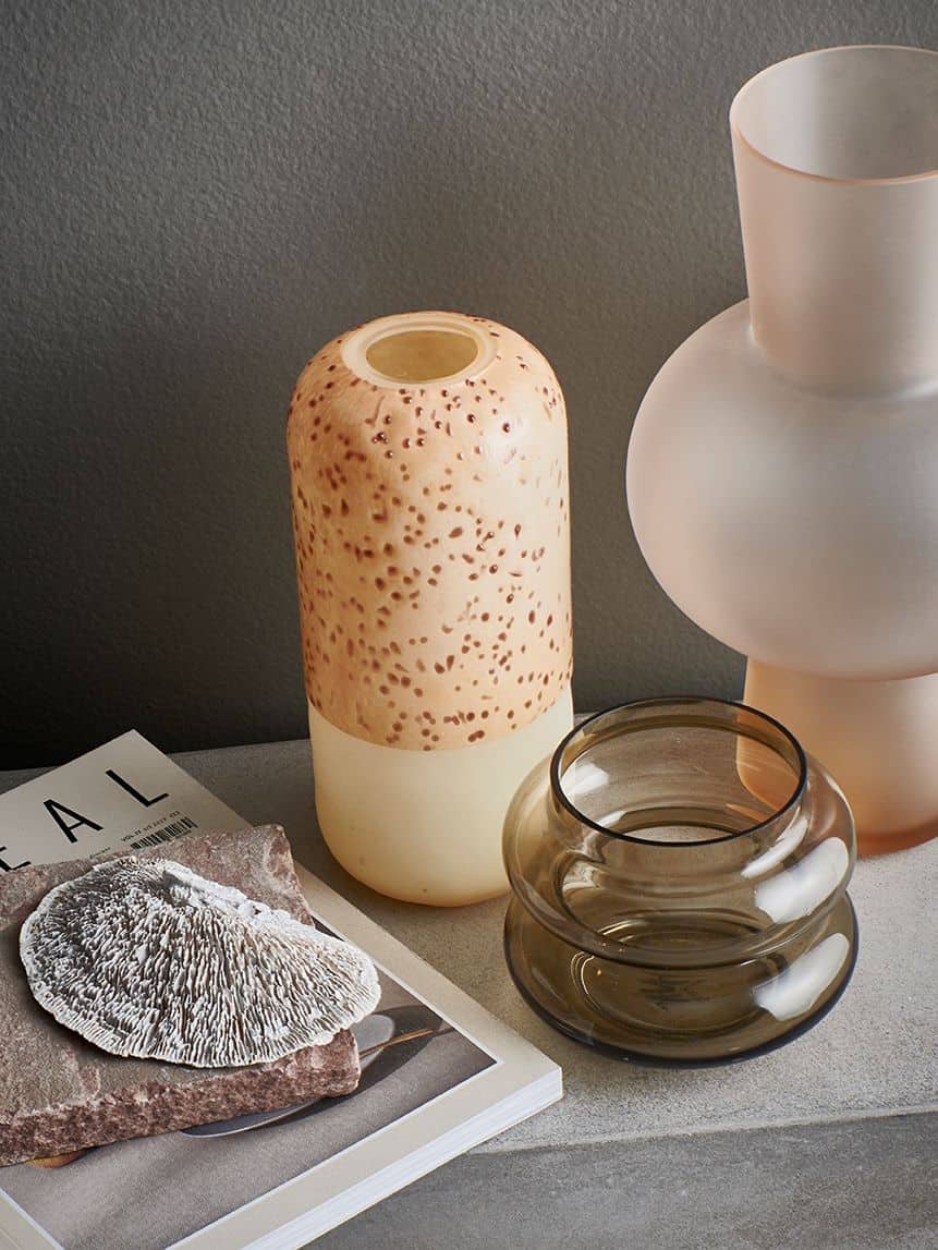 A minimalist still life displays the HKliving Glass Vase S, a frosted glass vase, and a double-walled smoky glass bowl—ideal for home decor—arranged next to a magazine featuring a mushroom image on a gray surface against a gray wall.