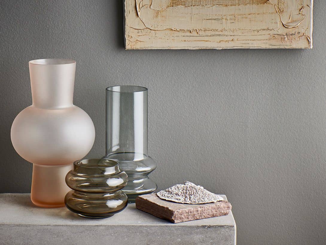The HKliving Glass Vase S by HKliving, in assorted shapes and colors, sits on a stone surface beside a textured rock beneath a beige abstract painting on a gray wall, creating striking minimalist home decor.