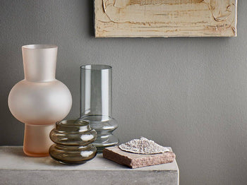 The HKliving Glass Vase S by HKliving, in assorted shapes and colors, sits on a stone surface beside a textured rock beneath a beige abstract painting on a gray wall, creating striking minimalist home decor.
