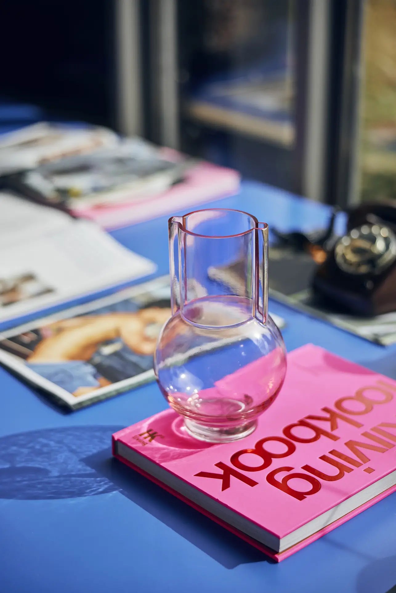The HKLIVING Glass Vase Sundae Pink, ideal for modern spaces, rests on a closed bright pink book on a blue table. Next to it are an open magazine and a vintage rotary phone, with sunlight streaming in and casting subtle shadows.