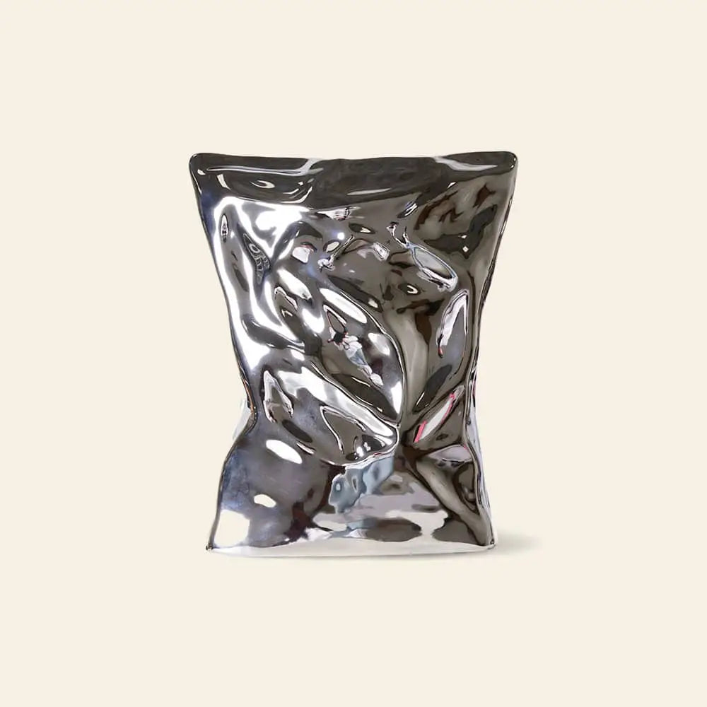 The HKLIVING HK Objects: Bag of Crisps Vase, a reflective metallic sculptural piece resembling a crumpled bag, stands upright against a plain beige background, adding playful decor from the HKLIVING brand.