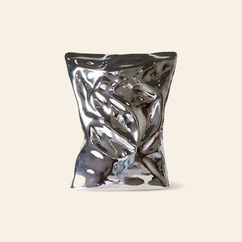 The HKLIVING HK Objects: Bag of Crisps Vase, a reflective metallic sculptural piece resembling a crumpled bag, stands upright against a plain beige background, adding playful decor from the HKLIVING brand.