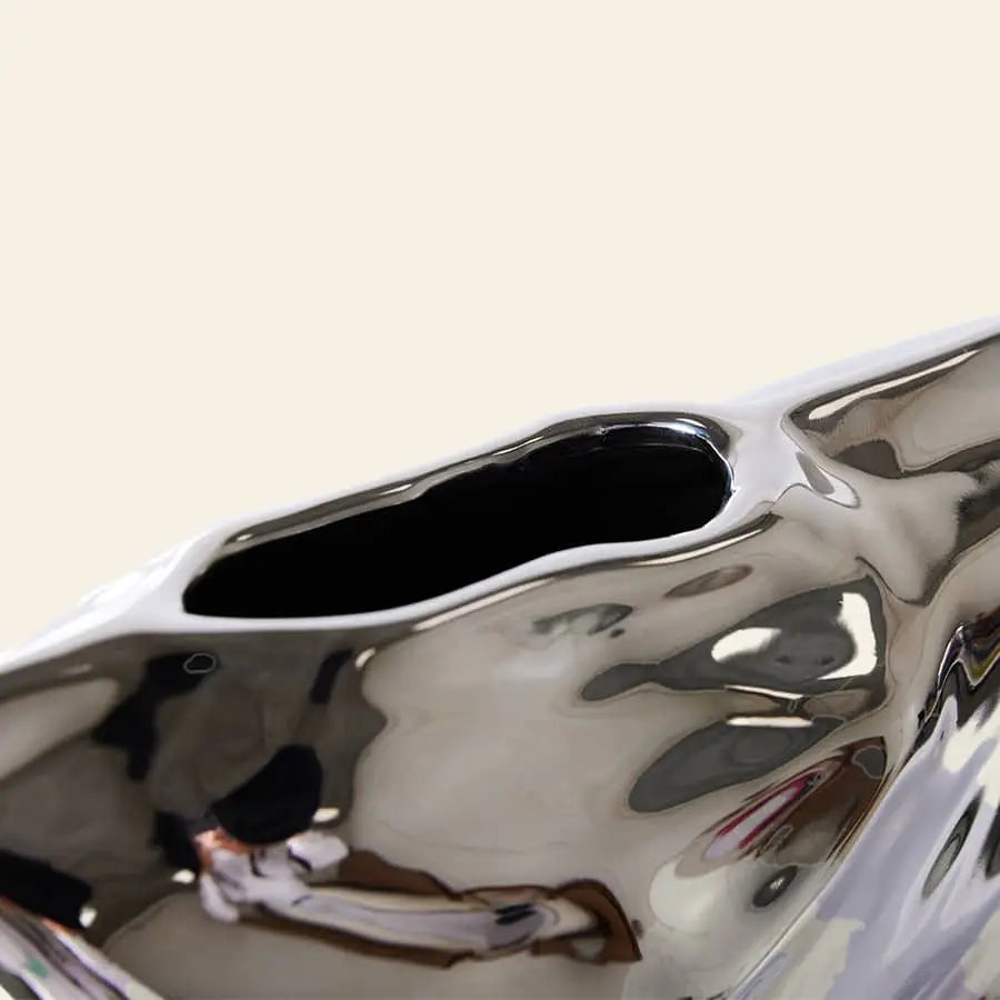 Close-up of the HKLIVING HK Objects: Bag of Crisps Vase by HKLIVING, featuring a shiny silver abstract design with a smooth, uneven surface and an irregular oval opening, perfect as playful decor against a light beige background.