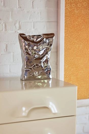 The HKLIVING HK Objects: Bag of Crisps Vase, with its shiny crumpled texture, brings playful style to a glossy cream dresser near a white brick wall and framed orange art—an ideal decor accent from HKLIVING.