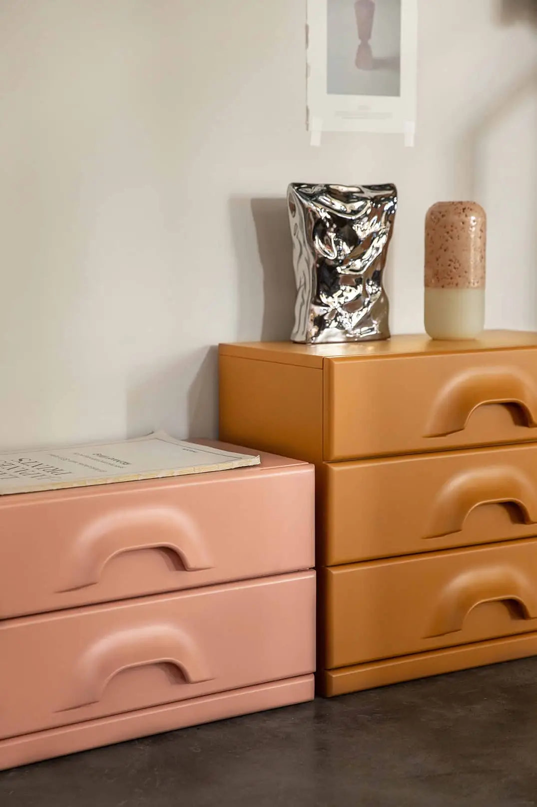 A pink and a mustard yellow drawer stand side by side against the wall, topped with a book, the HKLIVING HK Objects: Bag of Crisps Vase, a speckled beige jar, and an art print above—creating a playful display.