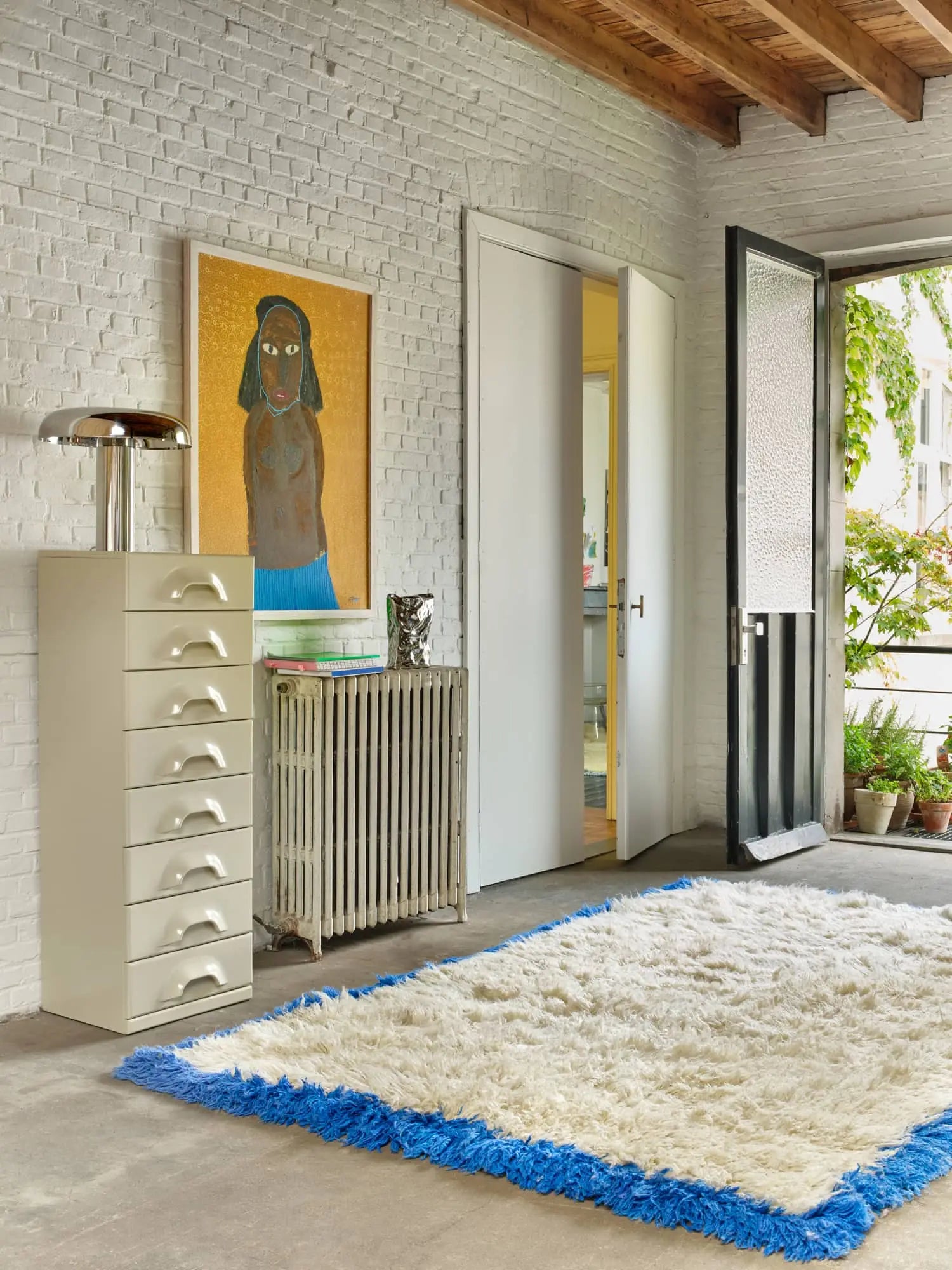 A bright room with white brick walls, playful decor including the HKLIVING HK Objects: Bag of Crisps Vase, a white shag rug with a blue border, stacked-drawer cabinet, woman’s artwork, open black door, radiator, and potted plants outside.