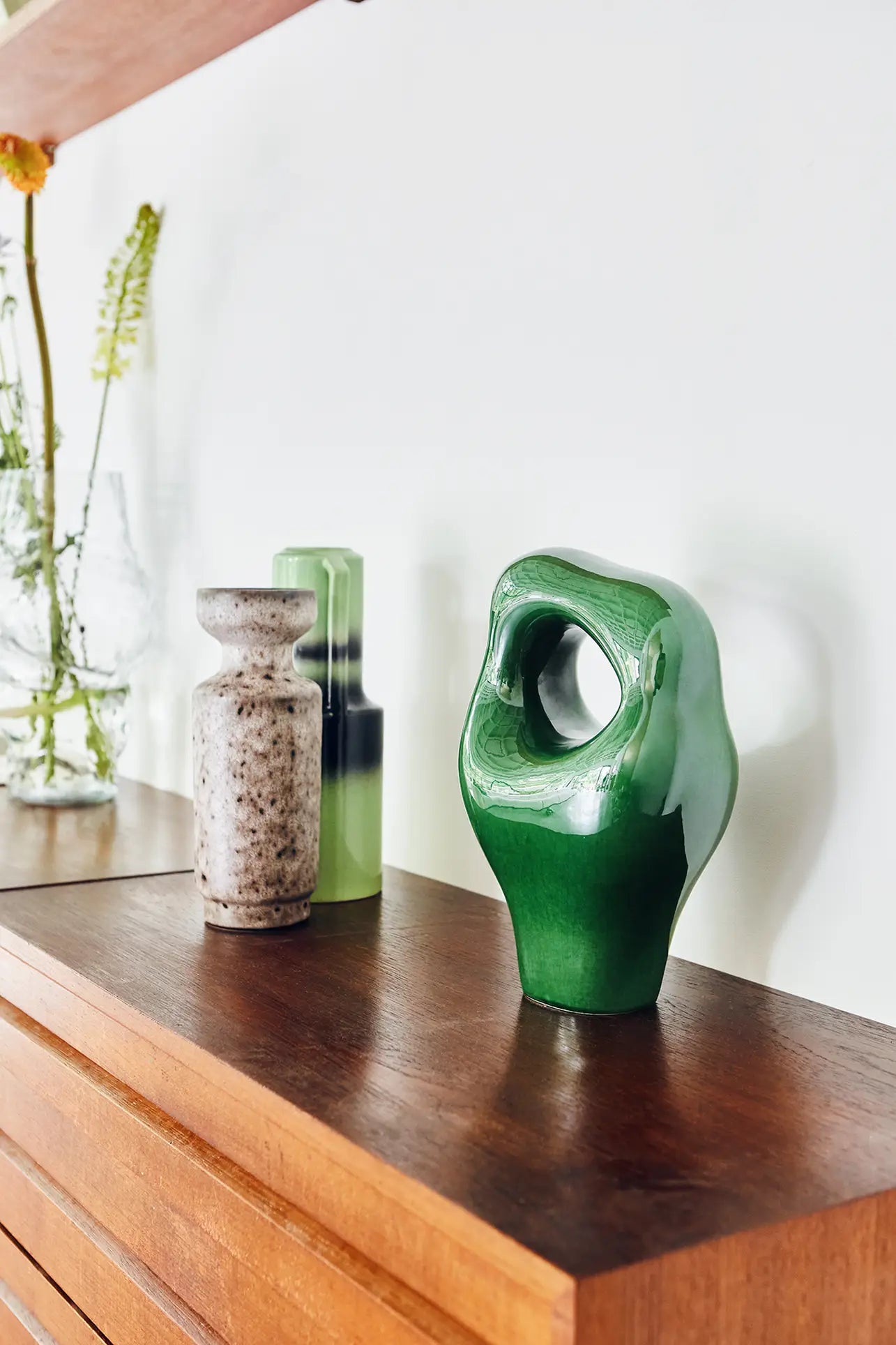 Three HKLIVING HK Objects ceramic sculptures—a speckled beige, a translucent green rectangular, and a glossy green abstract piece—sit on wood, perfect for modern decor. Blurred glass vases with flowers appear in the background.