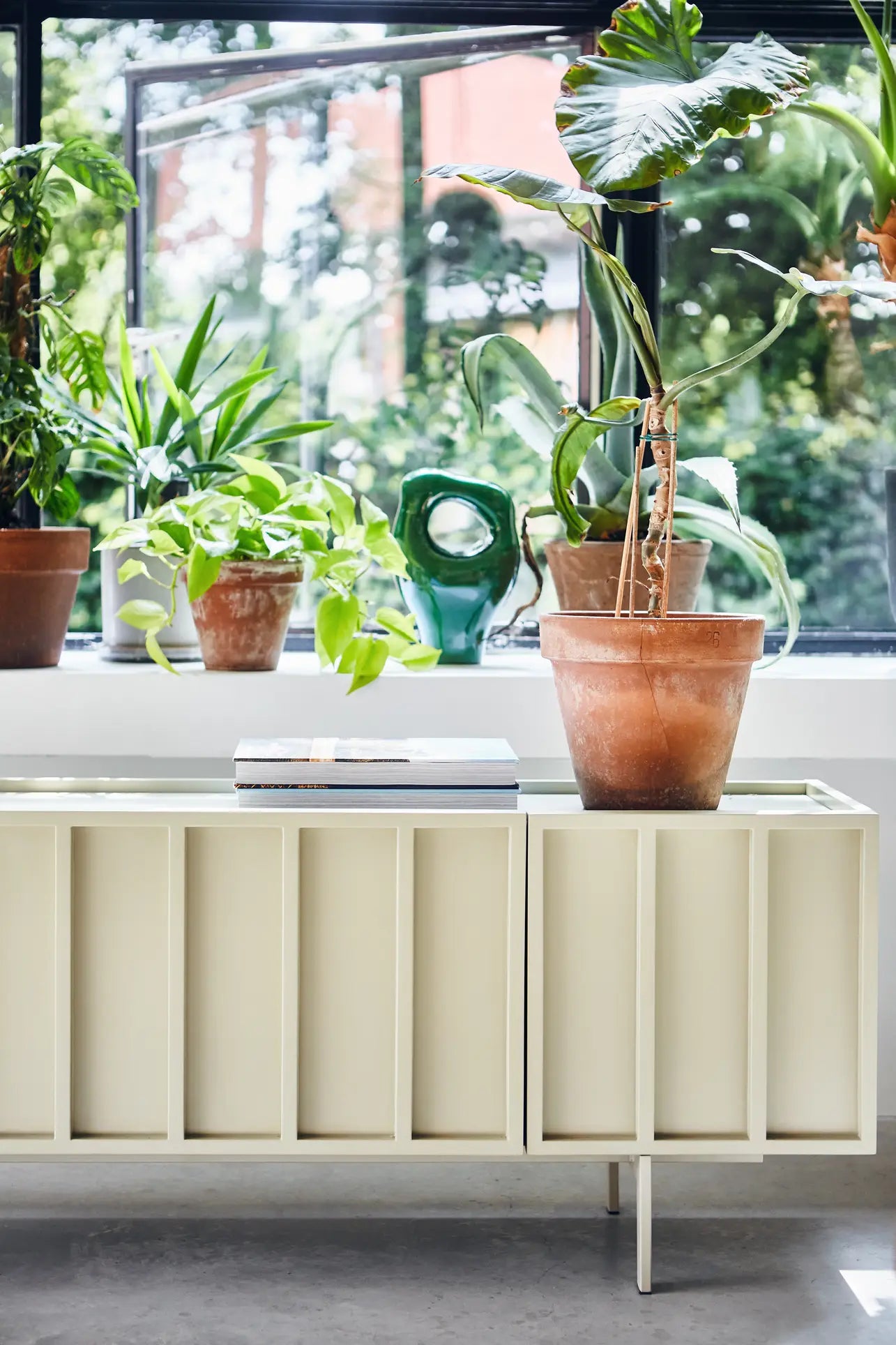 A modern white sideboard beneath a large window holds green plants, home decor, two stacked books, and the HKLIVING HK Objects Ceramic Sculpture by HKLIVING, with sunlight streaming in and trees visible outside.