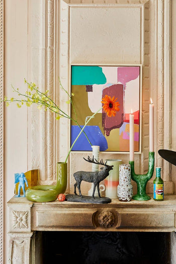 A mantel featuring eclectic decor: the HKliving HK Objects Ceramic Twisted Vase by HKliving, yellow flowers, a painting, sunflower, candles, a blue elephant figurine, deer sculpture, stoneware pieces, and colorful accents.